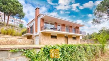 Buitenaanzicht van Villa Lloret met een huis van twee verdiepingen, voorzien van een stenen benedenverdieping, een bakstenen bovenverdieping en een breed balkonterras rondom, onder een bewolkte blauwe lucht.