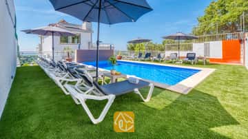 Privézwembad bij Villa Sandy Beach met rijen witte ligbedden, blauwe parasols en een groen kunstgrasveld onder een strakblauwe lucht.