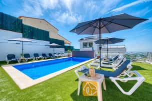 Privézwembad bij Villa Sandy Beach, omringd door kunstgras, grijze ligbedden met witte frames en grote parasols onder een strakblauwe lucht.