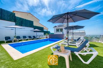 Privézwembad bij Villa Sandy Beach, omringd door kunstgras, grijze ligbedden met witte frames en grote parasols onder een strakblauwe lucht.