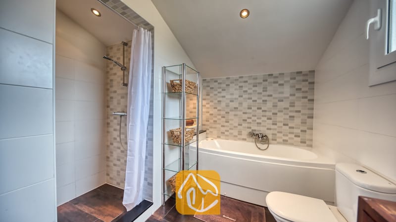 Moderne badkamer met een betegelde inloopdouche met een wit douchegordijn, een wit ligbad, een glazen wandrek met geweven manden en een toilet.