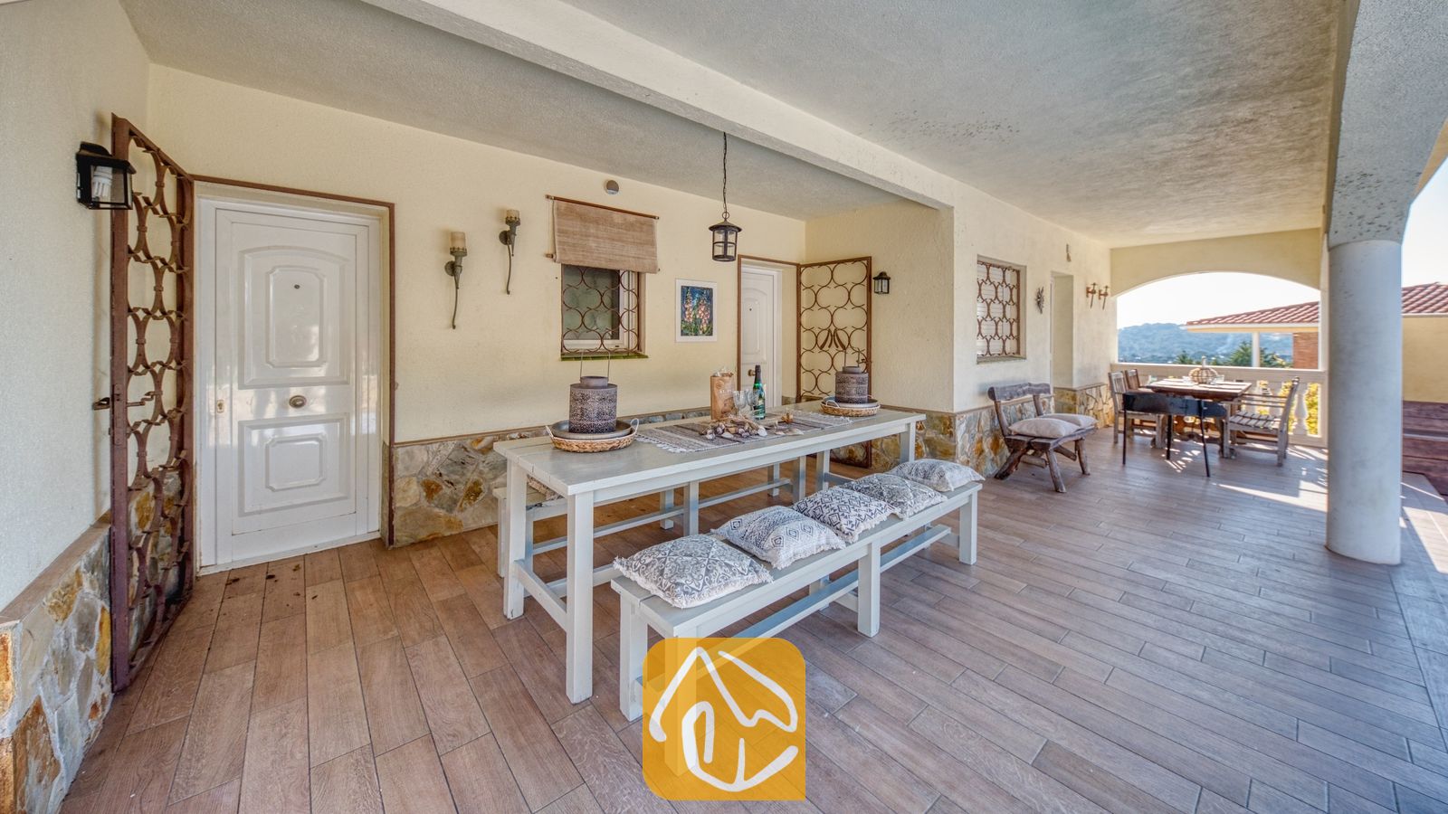 Overdekt buitenterras bij Villa Madonna met een lange witte eettafel met bijpassende banken, kussens met een patroon, stenen funderingsmuren en een tegelvloer.