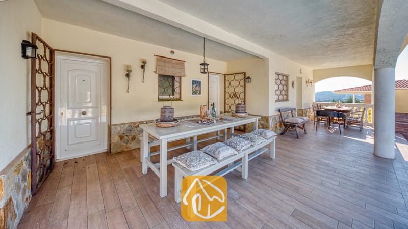 Overdekt buitenterras bij Villa Madonna met een lange witte eettafel met bijpassende banken, kussens met een patroon, stenen funderingsmuren en een tegelvloer.