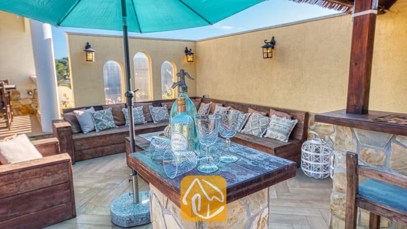Buitenbar bij Villa Madonna met een stenen barblad, glaswerk, flessen, een grote blauwgroene parasol en houten banken met gedessineerde kussens.