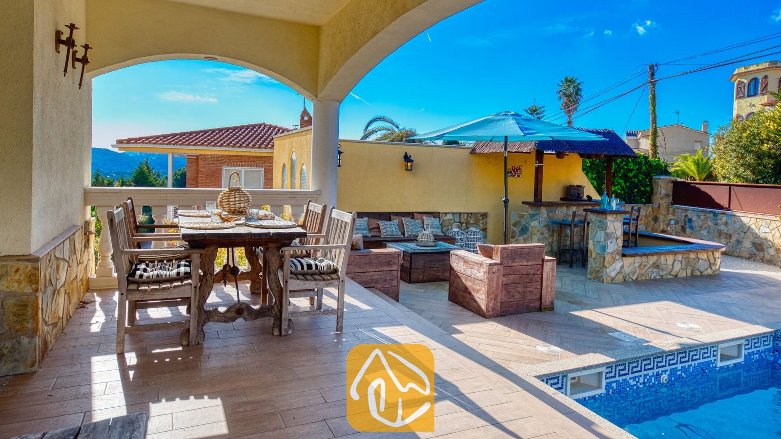 Overdekt terras bij Villa Madonna met een rustieke houten eettafel, fauteuils en een betegeld buitenzwembad met een blauwe parasol.