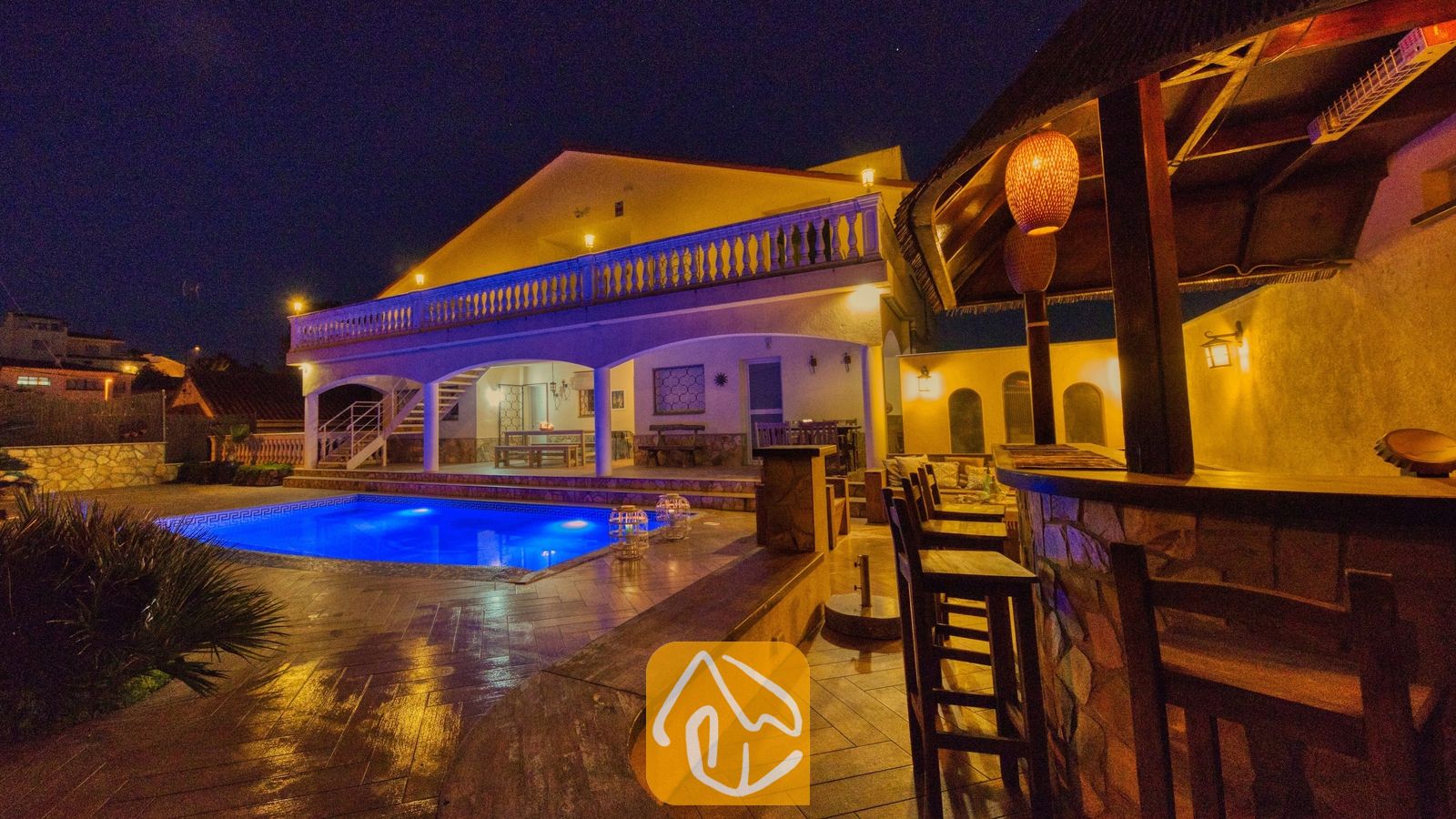 De buitenzijde van Villa Madonna bij nacht, met een verlicht zwembad, een overdekte buitenbar met houten krukken en een verlicht terras met een balustrade.