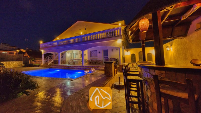 De buitenzijde van Villa Madonna bij nacht, met een verlicht zwembad, een overdekte buitenbar met houten krukken en een verlicht terras met een balustrade.