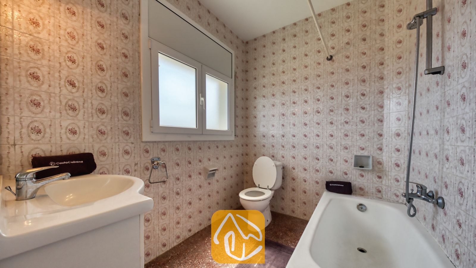 Badkamer voorzien van muren met patroon van keramische tegels, een witte wastafel, een ligbad met een verticale douchekop en een toilet onder een matglas raam.
