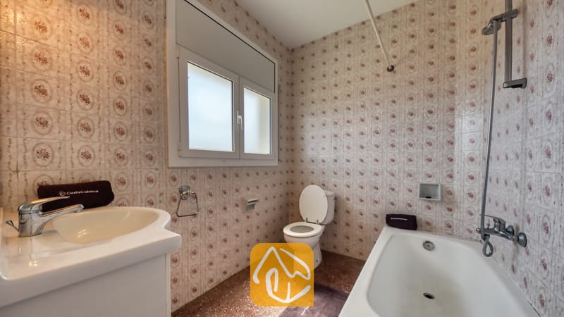 Badkamer voorzien van muren met patroon van keramische tegels, een witte wastafel, een ligbad met een verticale douchekop en een toilet onder een matglas raam.