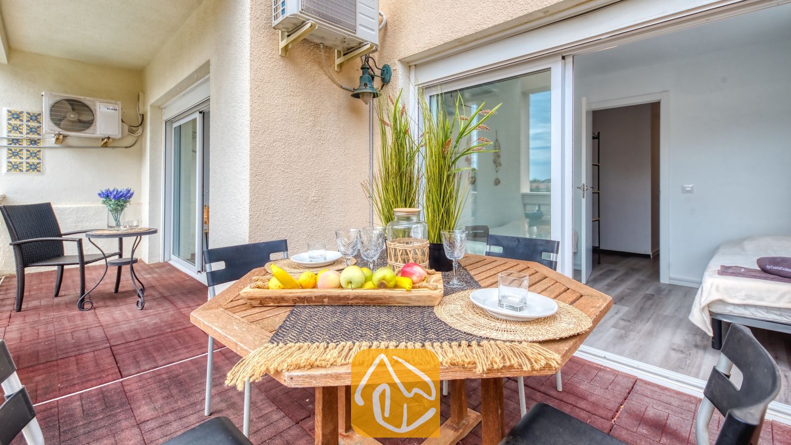 Houten eettafel voor buiten op een balkon met een schaal fruit, glazen en een pot met siergras, buiten Villa Madonna.