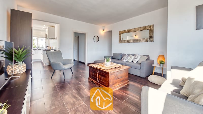 Woonkamer van Villa Madonna met een grijze bank, een salontafel van donkerhout, een fauteuil en tegelvloeren, met uitzicht op de keuken.