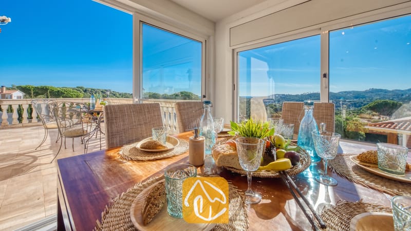 Eettafel binnen in Villa Madonna, gedekt met gevlochten placemats, glaswerk, brood en fruit, omringd door grote glazen ramen met uitzicht op een terras en een groen landschap.