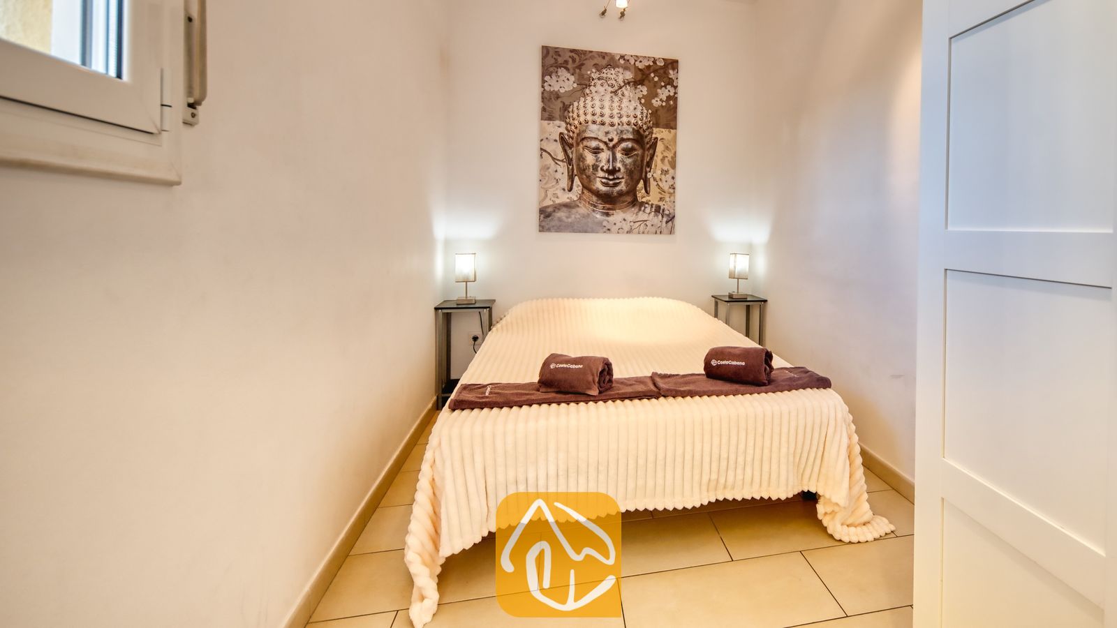 Slaapkamer met een tweepersoonsbed voorzien van een lichtkleurige, geribbelde sprei, opgerolde bruine handdoeken en een groot wandkleed met een Boeddha-afbeelding erboven.