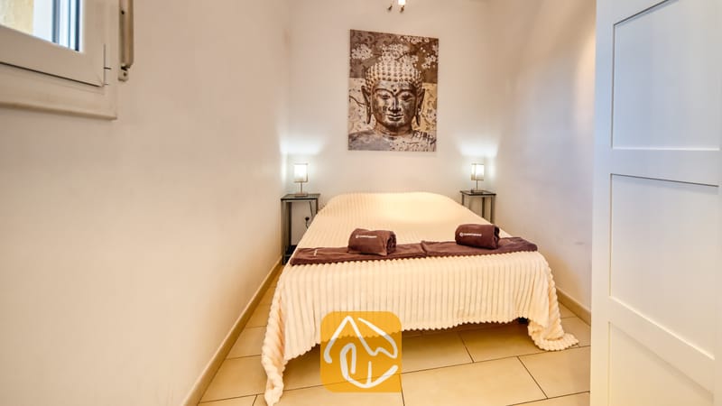 Slaapkamer met een tweepersoonsbed voorzien van een lichtkleurige, geribbelde sprei, opgerolde bruine handdoeken en een groot wandkleed met een Boeddha-afbeelding erboven.