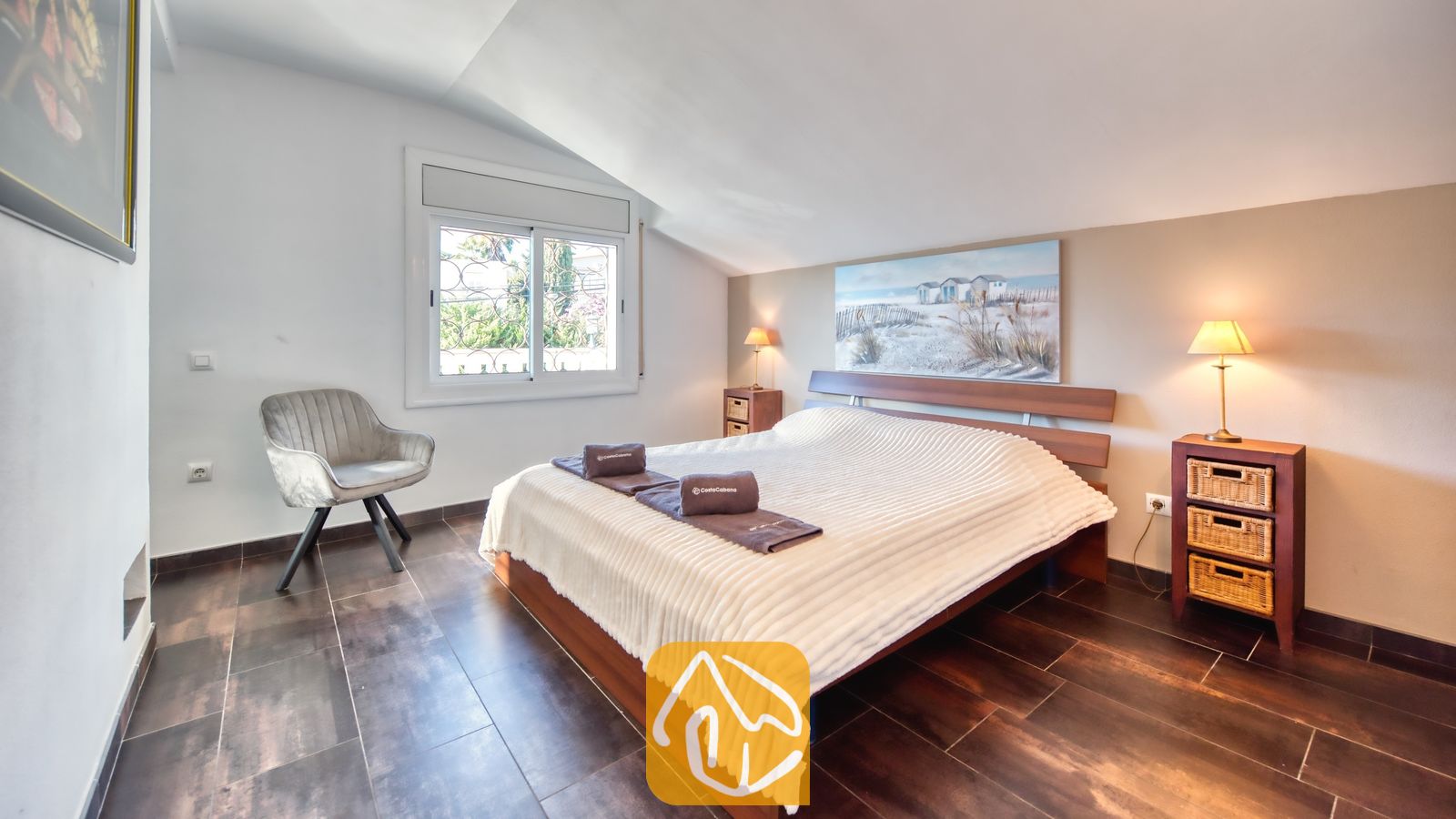 Slaapkamer met een houten bedframe, een wit structuurdekbed, twee nachtkastjes met rieten lades en een grijze fauteuil bij het raam.