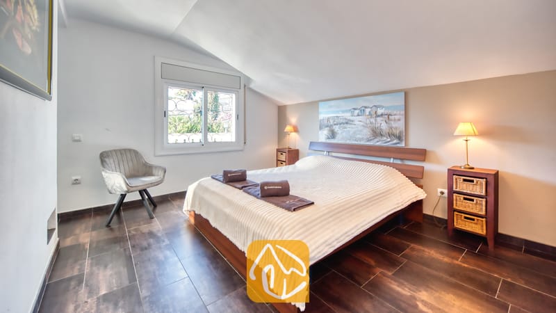 Slaapkamer met een houten bedframe, een wit structuurdekbed, twee nachtkastjes met rieten lades en een grijze fauteuil bij het raam.