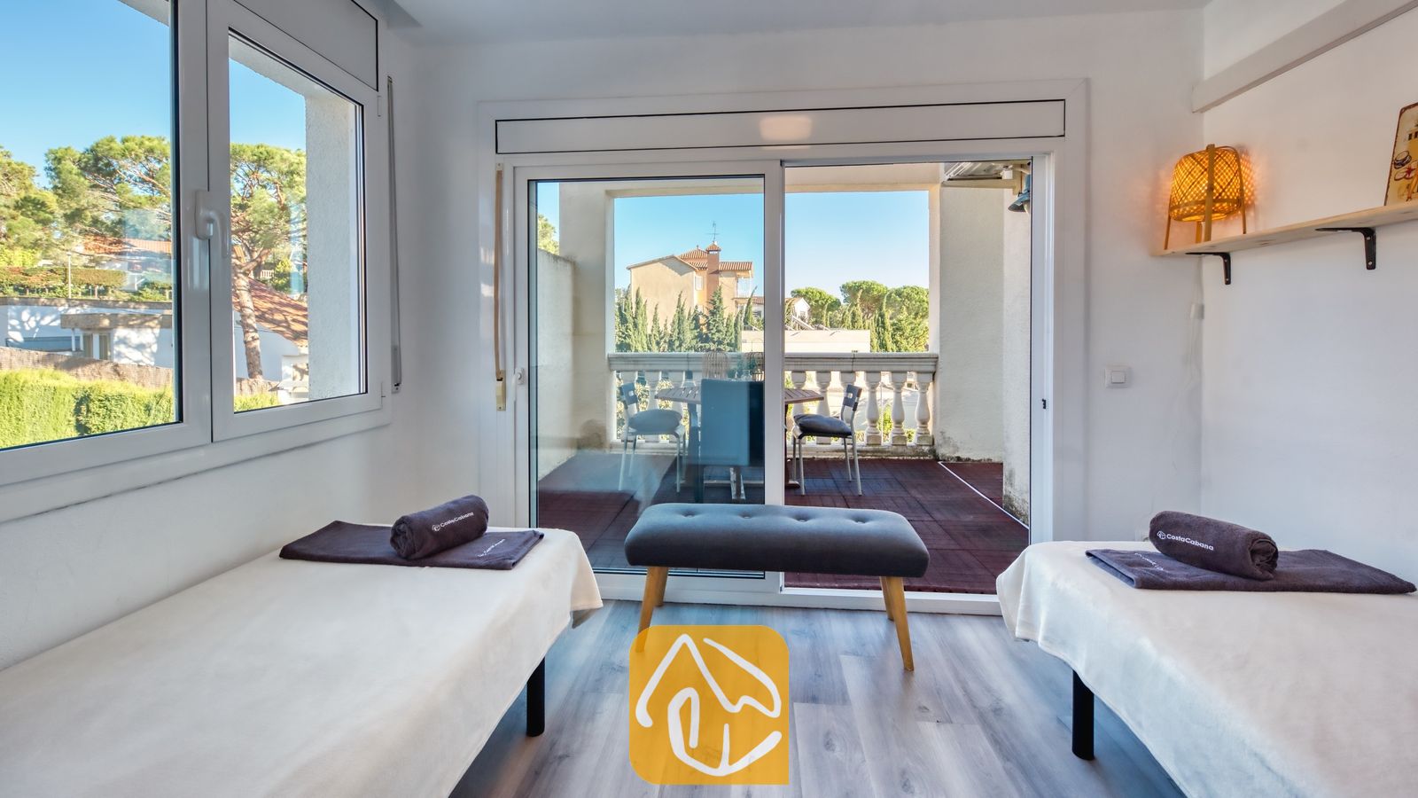 Tweepersoonskamer met twee aparte bedden in Villa Madonna, voorzien van donkergrijze opgerolde handdoeken, een houten bankje en schuifpui naar een balkon.