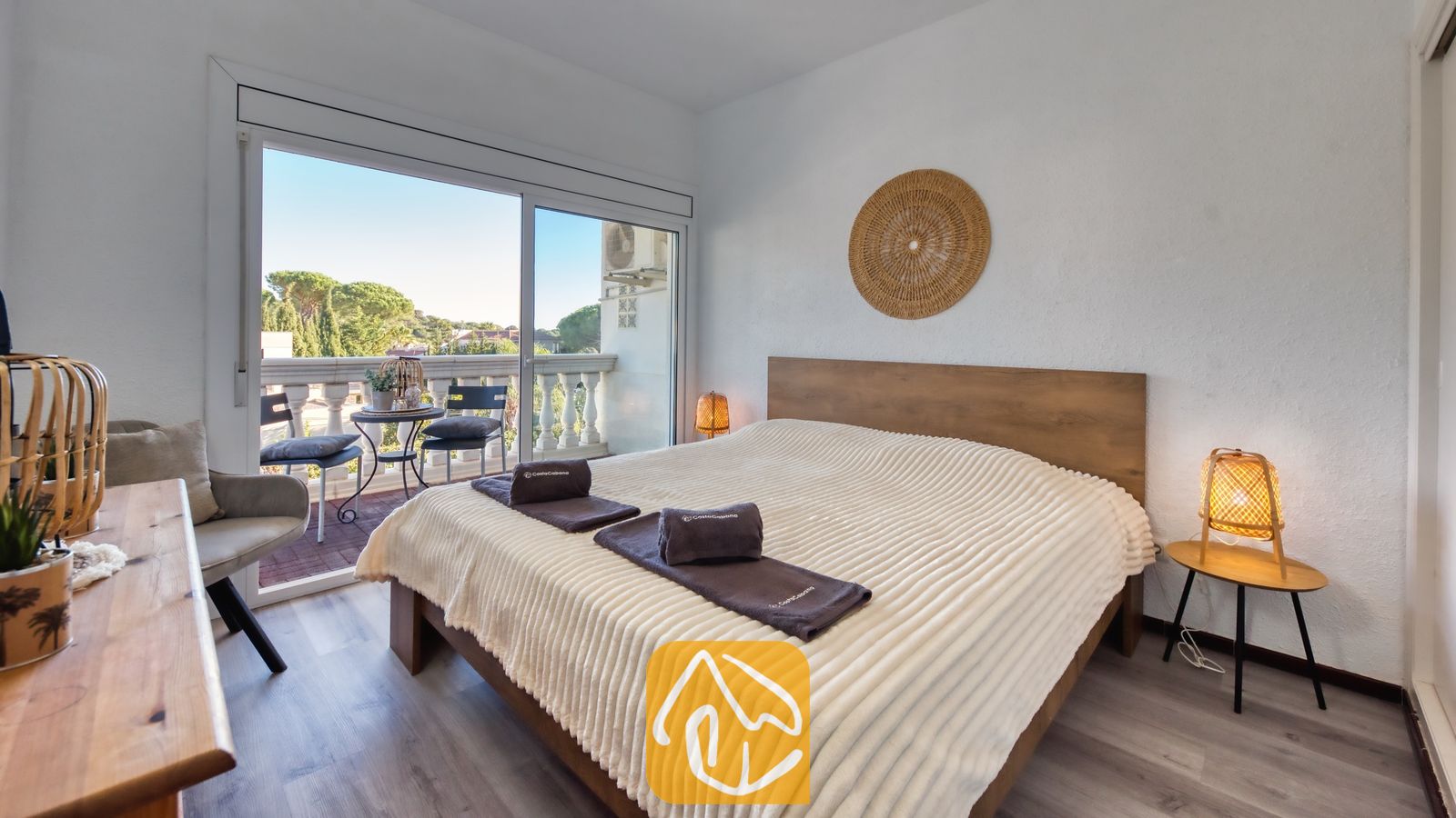Slaapkamer in Villa Madonna met een houten bed voorzien van een crèmekleurige, geribbelde sprei, geweven bedlampjes en een schuifpui die uitkomt op een balkon.