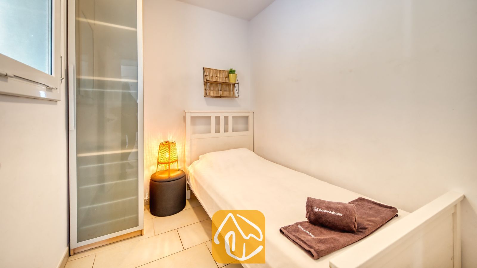 Slaapkamer met een wit houten bed, bruine opgevouwen handdoeken, een geweven bedlampje op een donkere kruk en een hoge kledingkast van matglas.