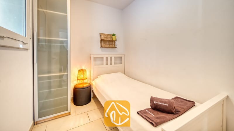 Slaapkamer met een wit houten bed, bruine opgevouwen handdoeken, een geweven bedlampje op een donkere kruk en een hoge kledingkast van matglas.