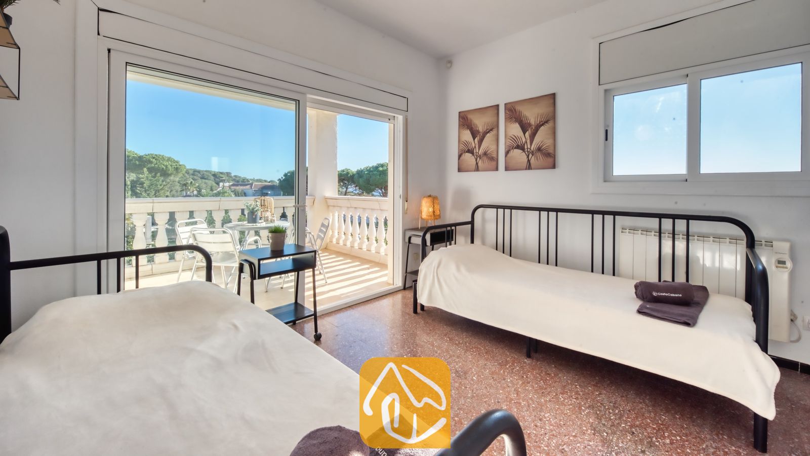 Tweepersoonskamer met twee eenpersoonsbedden met een zwart metalen frame, wit beddengoed en een schuifpui die uitkomt op een zonnig balkon bij Villa Madonna.
