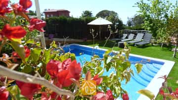 Het privézwembad bij Villa Noa, omlijst door rode bougainvillea, met ligstoelen en een witte parasol op het gazon op de achtergrond.