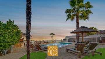 Privé buitenzwembad bij Villa Tropicana, omringd door houten ligstoelen, palmbomen en rieten parasols onder een avondhemel.