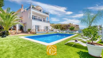 Privézwembad en witte ligbedden op een groen gazon bij Villa Maribel in Spanje, met een witte villa met meerdere verdiepingen, balkons en palmbomen.