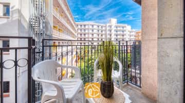 Balkon van Appartement Gaudi met twee witte kunststoftoelen, een klein tafeltje met een plant in pot en een metalen balustrade die uitkijkt over de naburige witte gebouwen.